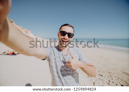 Happy young man dressed in t-shirt taking a selfie while standing at the beach and showing thumbs up