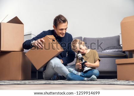 Picture of happy man and small boy with microscope sitting on floor among cardboard boxes