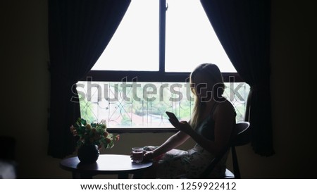 silhouette. girl sits on a chair against the window, uses her phone and drinks water from a glass