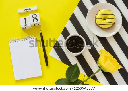 Wooden cubes calendar April 18th. Cup of coffee, yellow donut and rose on black and white napkin, empty open notepad for text on yellow background. Concept stylish workplace Top view Flat lay Mockup