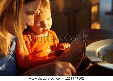 mom trying to feed toddler, while he watches cartoons via smartphone during a sunset in  a restaurant 