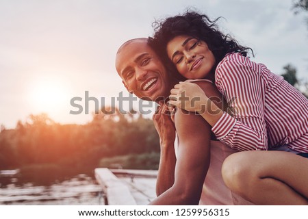 Happy moments together. Happy young couple embracing and smiling while sitting on the pier near the lake Royalty-Free Stock Photo #1259956315