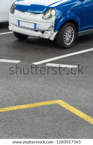 Damaged blue and white little car with a crash on her frontal bumper, parked in an outside parking with white and yellow lines on the asphalt by day.