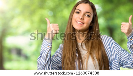 Young girl with striped shirt giving a thumbs up gesture with both hands and smiling at outdoors