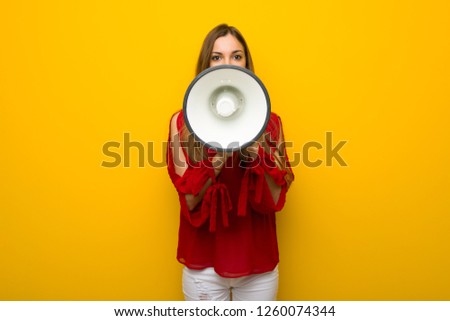 Young girl with red dress over yellow wall shouting through a megaphone to announce something