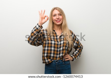 Blonde young girl over white wall showing an ok sign with fingers