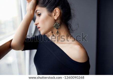 Beautiful brunette in a black dress stands near the window                               