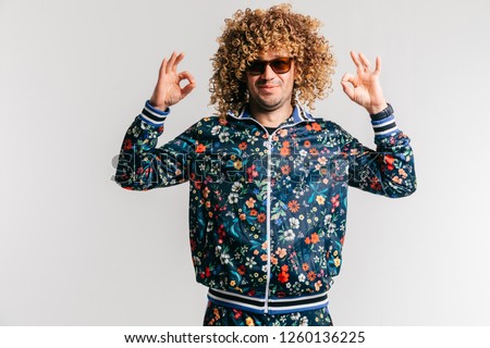 Adult positive smiling funky man with afro curly hair style in suglasses, multicolored clothes making okay gesture with hands on white studio background. Funny portrait of artistic stylish male person