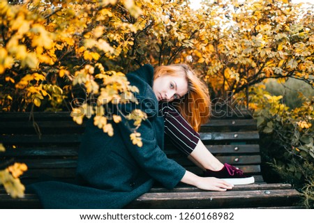 Young beautiful redhead girl sitting on bench under dog rose bushes. Cute female lifestysle soft focus portrait. Pensive blue eyed teen with thoughtful calm face posing outdoor. Cute stylish woman