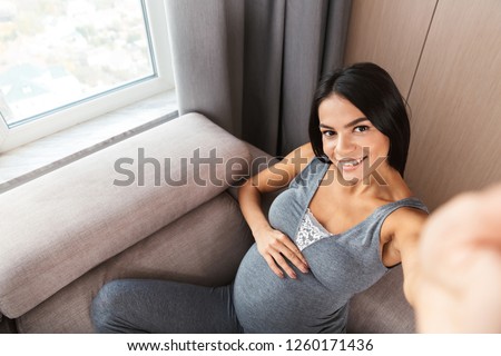 Happy young pregnant woman sitting on a couch at home, taking a selfie