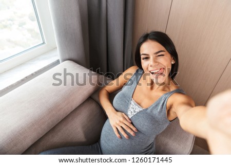 Happy young pregnant woman sitting on a couch at home, taking a selfie