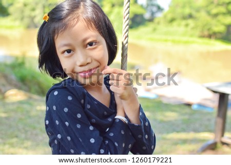 Happy kid female smile and make finger  show sign of mini heart, girl play swing around nature outdoor near river and green environment, International Children's day, Valentine's  day concept 