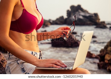 Lady freelancer in red bikini swimsuit with a laptop remote work on the beach on the sunset.freelancing girl working near the rocks and the sea.business women on vacation in a tropical desert paradise