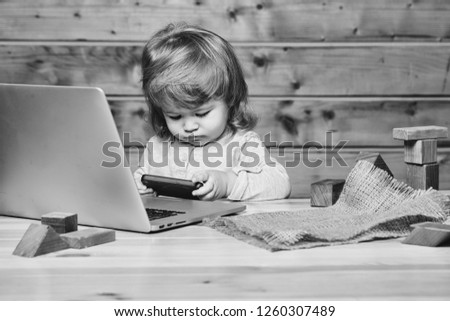 Cute funny little baby boy with long blonde curly hair playing on computer and mobile phone near toy building blocks indoor on wooden background, horizontal picture