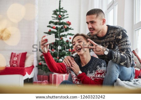 Taking a selfie. Guy showing gesture of two fingers. Happy young people sits on the windowsill in the room with christmas decorations.