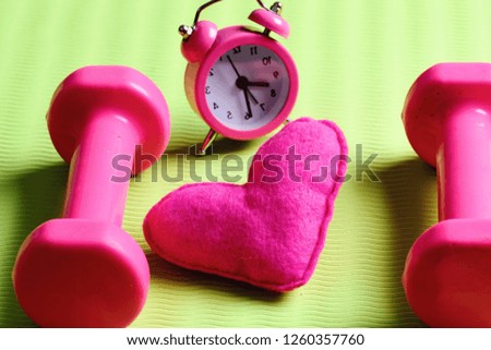Morning workout concept. Dumbbells in pink color next to alarm clock and soft toy heart on green background. Love of sports and early training. Heart decoration near plastic barbells on yoga mat