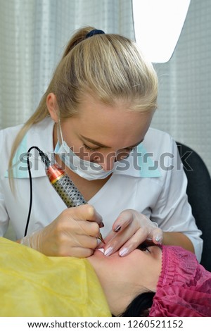 Master beautician, blonde, bent over lying European girl and makes her permanent lip makeup in the beauty salon. Close up. The view from the top. Vertical orientation of the image