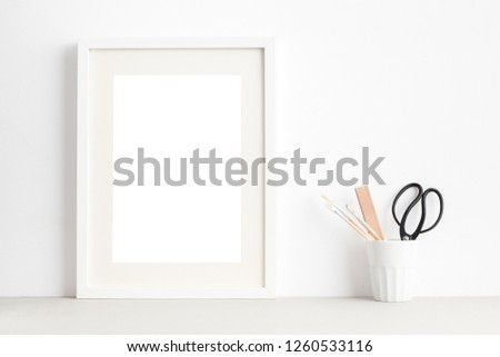 White poster frame mock up and artist tools in a mug, against white wall.