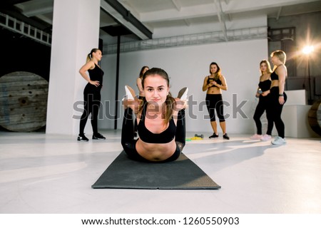 Young smiling woman warming up arching stretching their back holding legs and working out in a gym yoga class. Attractive young sport girls are doing yoga together on the background.