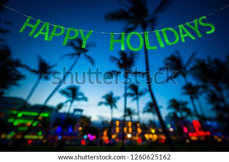 Happy Holidays message hanging in glittery party banner above tropical palm trees of the glowing neon lights of the skyline of Miami Beach, Florida, USA