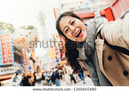 young female traveler cheerfully taking selfie on busy street in tsutenkaku osaka japan. girl smiling at the camera self portrait with peffer fish balloon floating on the sky in sunshine.