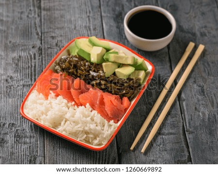 A plate of Hawaiian rice, avocado, salmon and kelp and soy sauce on a black rustic table. The view from the top.