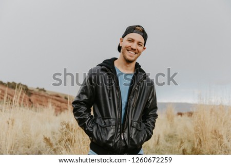 Portrait of Young Good Looking Handsome Man Smiling with Backwards Hat in Leather Jacket Outside in Isolated Rural Field of Tall Grass in the Meadow Nature Outside Autumn Season Royalty-Free Stock Photo #1260672259