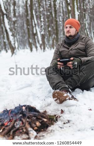a man uses a mobile phone, sitting by the fire in a snowy forest. Mobile networks. Call for help. battery discharge and signal loss, smartphone for tourism and trips, survival, vertical photo