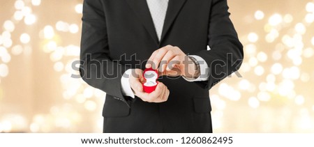 valentines day, proposal and engagement concept - close up of man with diamond ring in little red gift box over festive lights background