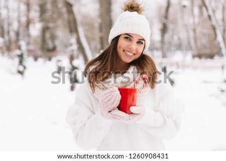 Woman holding winter cup close up on park. Woman hands in gloves holding a cozy mug with hot cocoa, tea or coffee and a candy cane. Winter and Christmas time concept.