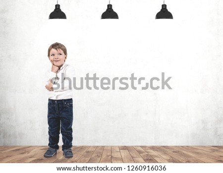 Portrait of adorable little boy with fair hair wearing blue jeans and white shirt standing near concrete wall and thinking. Mock up