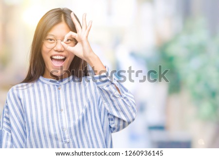 Young asian woman over isolated background doing ok gesture with hand smiling, eye looking through fingers with happy face.