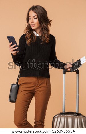 Image of cheerful woman 20s with baggage holding mobile phone and travel ticket isolated over beige background