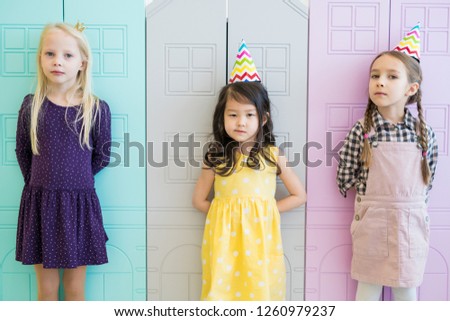 Serious confident attractive girls in stylish clothing standing against cartoon houses and leaning on them while looking at camera