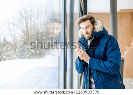 Man dressed in winter clothes feeling cold standing near the window at home with no heating