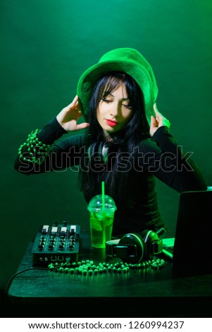 Portrait of DJ young irish girl on green background. DJ woman with headphne and green hat plays music on the party of St. Patrick.