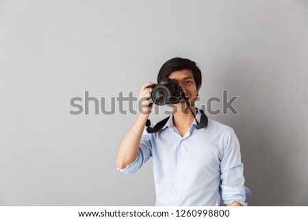 Happy asian man standing isolated over gray background, taking a picture with photo camera