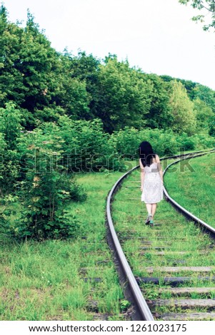 Girl walking away on railway near green forest. Walks to wonderland. Old abandoned railroad with young woman. Summer surreal landscape with woods  and girls long alone journey concept, pastel colors