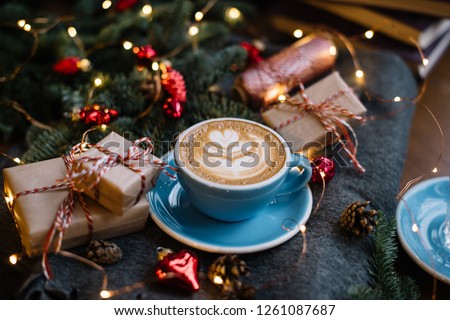 Delicious fresh festive morning cappuccino coffee in a ceramic blue cup on the warm cover with little wrapped gifts, red ornamentals, fireflies and spruce branches Royalty-Free Stock Photo #1261087687