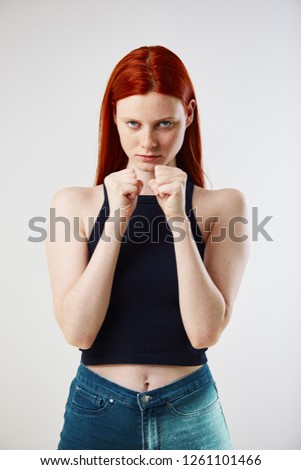 Charming redhead long-haired girl dressed in black top and jeans keeps hands in fists on the white background in the studio