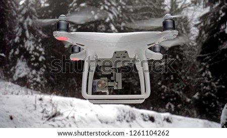 Close up photo of a hovering drone. The Aerial UAV is pictured in a winter, snowy environment in front of some snow covered alpine trees.