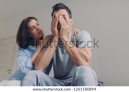 Photo of depressed male dressed casually, his beautiful woman comforts him, sit together in bedroom, have problems in family life. Affectionate wife consoles husband in difficult situation. Royalty-Free Stock Photo #1261180894