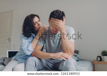 Attractive female consoles her sad boyfriend who has depression and some problems, pose at bedroom on bed, sit against domestic interior. Wife and husband try solve difficult situation in life. Royalty-Free Stock Photo #1261180915