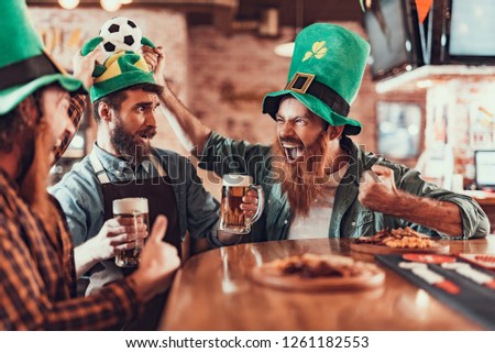 Young bearded bartender with beer standing near bar counter while two men holding his hat with soccer ball. They screaming with joy and wearing traditional green headgears with shamrock