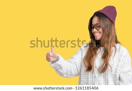 Young beautiful brunette hipster woman wearing glasses and winter hat over isolated background Looking proud, smiling doing thumbs up gesture to the side