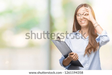 Young asian business woman holding clipboard over isolated background with happy face smiling doing ok sign with hand on eye looking through fingers