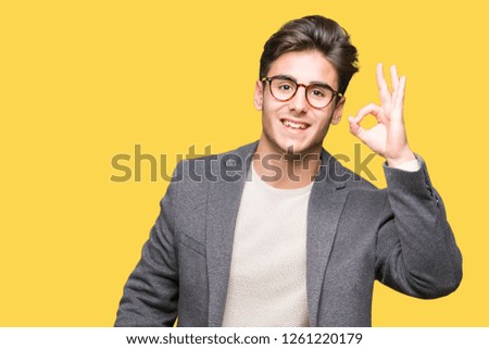 Young business man wearing glasses over isolated background smiling positive doing ok sign with hand and fingers. Successful expression.