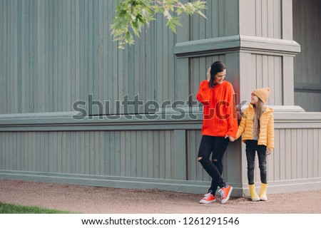 Horizontal view of fashionable European female in red loose sweater, ripped jeans, holds hands together with her younger sister, pose outside together, have walk in open air, have friendly relations