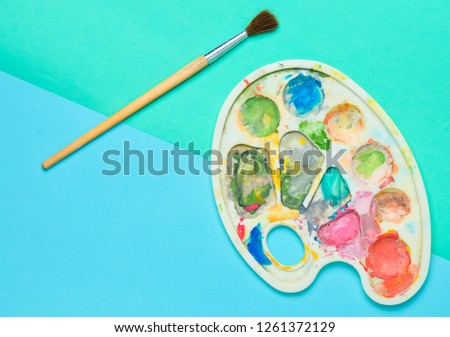 Plastic palette with gouache paint and brush on a colored paper background. Top view.