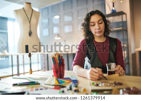 Portrait of creative young woman drawing sketches for jewelry while working at table in sunlit shop, copy space